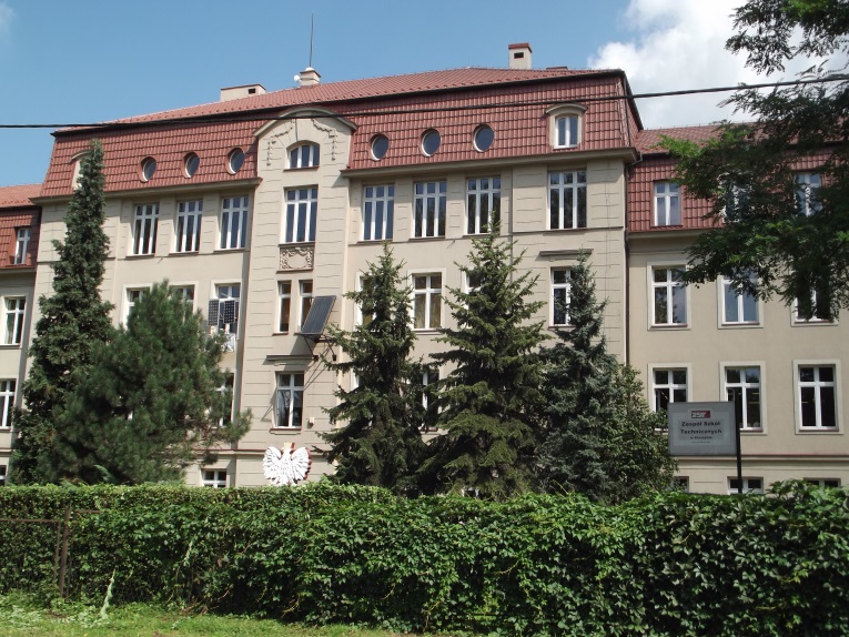 Czterokondygnacyjny budynek o jasnej elewacji i czerwonym dachu. Fasada z symetrycznym układem okien. Przed budynkiem drzewa iglaste i żywopłot. Po prawej stronie tablica z napisem 'Zespół Szkół Technicznych".